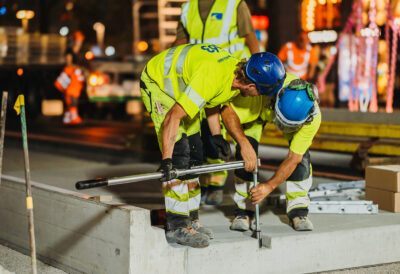 Zwei Bauarbeiter in Warnkleidung verschrauben bei Nachtarbeiten präzise ein vorgefertigtes Beton-Bustrassenelement.