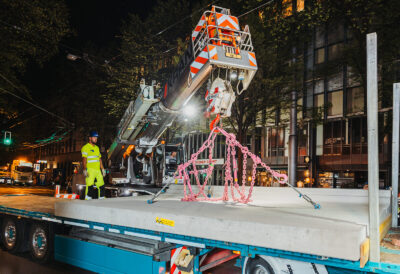 Ein Mobilkran hebt eine schwere, vorgefertigte Beton-Busfahrbahnplatte vom Lkw-Tieflader auf einer nächtlichen Baustelle.