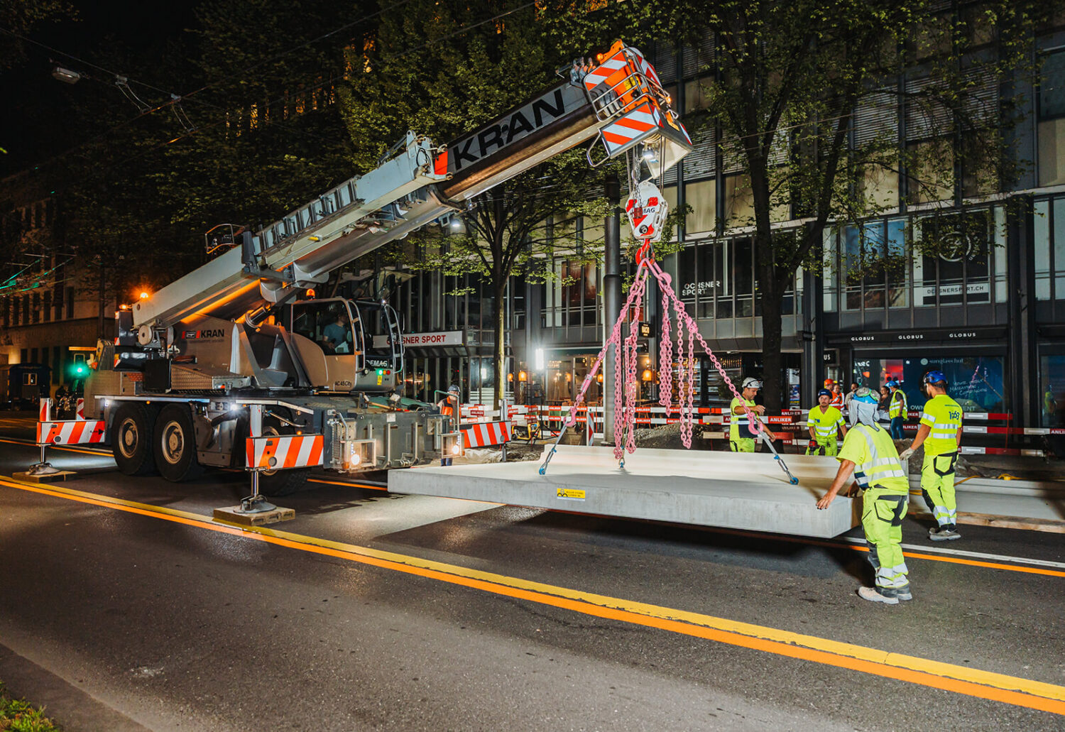 Bauarbeiter führen eine schwebende, vorgefertigte Betonplatte am Mobilkran präzise zur Montageposition auf einer nächtlichen Strassenbaustelle.