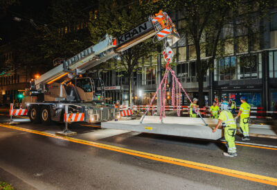 Bauarbeiter führen eine schwebende, vorgefertigte Betonplatte am Mobilkran präzise zur Montageposition auf einer nächtlichen Strassenbaustelle.